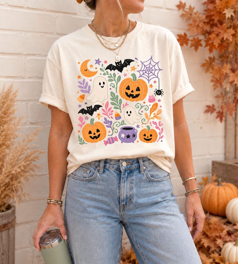 Może przedstawiać: Kremowy t-shirt z kr&oacute;tkim rękawem z kolorowym nadrukiem graficznym o tematyce Halloween, przedstawiającym pomarańczowe dynie, białe duszki, czarne nietoperze, fioletowy kociołek, pajęczynę, p&oacute;łksiężyc oraz r&oacute;żnego rodzaju cukierki.
