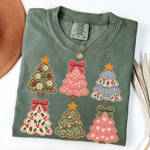 May include: A sage green short-sleeve t-shirt featuring a graphic print of six stylised Christmas trees in various colours and patterns, including floral, polka dot, and knit designs. The shirt has a Comfort Colors brand label at the neck.