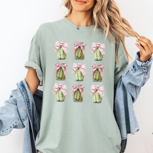 May include: A light green t-shirt with a repeating pattern of nine jars of pickles tied with pink bows. Each jar contains green pickles.