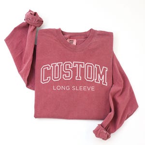 Op de afbeelding: Een bordeauxrode longsleeve met het woord "CUSTOM" in witte, collegiale letters. De woorden "LONG SLEEVE" staan eronder. De shirt heeft een ronde hals en opgerolde mouwen.