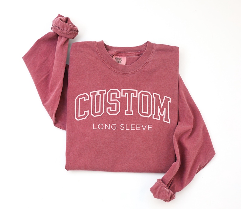 Op de afbeelding: Een bordeauxrode longsleeve met het woord "CUSTOM" in witte, collegiale letters. De woorden "LONG SLEEVE" staan eronder. De shirt heeft een ronde hals en opgerolde mouwen.