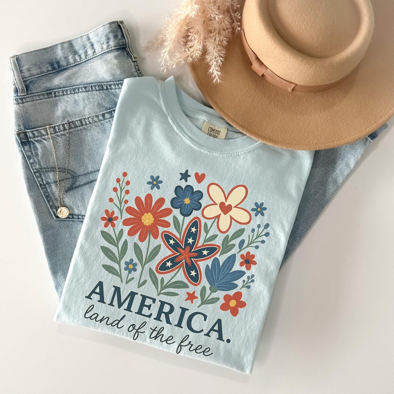 Peut inclure: Un t-shirt bleu clair avec un motif floral compos&eacute; de fleurs rouges, blanches et bleues. Le texte "AMERICA. land of the free" est imprim&eacute; sur le t-shirt.