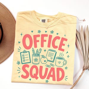 May include: A pale yellow t-shirt featuring the text OFFICE SQUAD in large pink letters. The graphic includes teal illustrations of a clipboard, a pencil holder, an apple, a notepad, a coffee mug, paperclips, small hearts, and flowers.