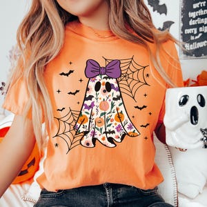 May include: Orange t-shirt with a floral ghost design, purple bow, bats, and spiderwebs. The ghost has a floral pattern with pumpkins and flowers. A Halloween-themed mug is held in the hand.