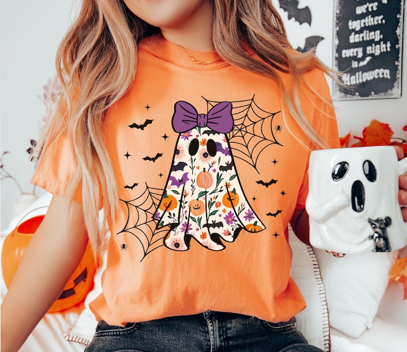 May include: Orange t-shirt with a floral ghost design, purple bow, bats, and spiderwebs. The ghost has a floral pattern with pumpkins and flowers. A Halloween-themed mug is held in the hand.