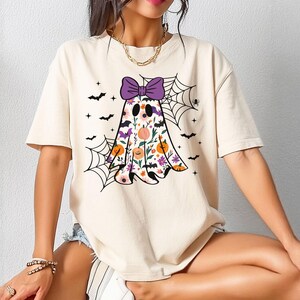 May include: Cream-coloured t-shirt featuring a Halloween ghost design. The ghost is adorned with a purple bow, floral patterns, and a pumpkin. Black bats and spiderwebs complete the festive design.