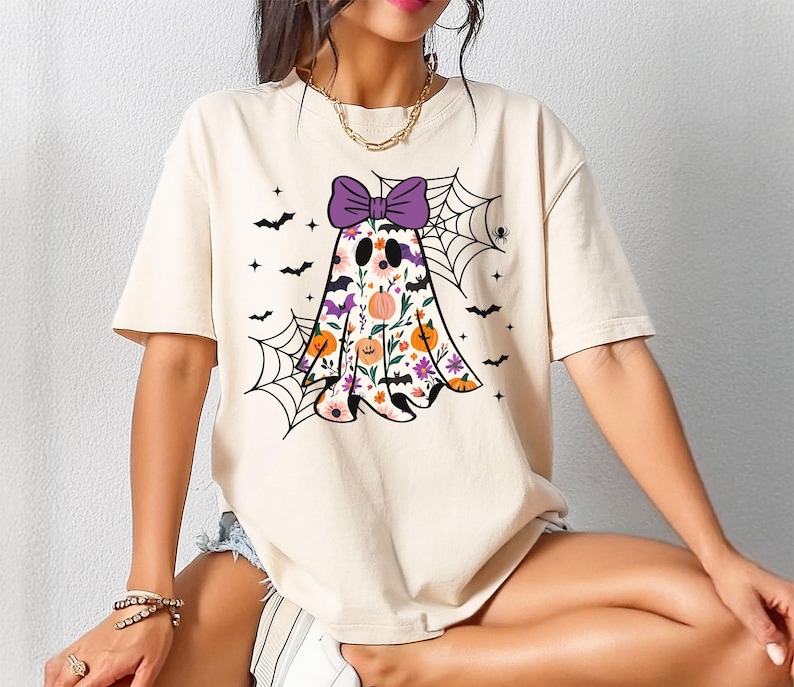 May include: Cream-coloured t-shirt featuring a Halloween ghost design. The ghost is adorned with a purple bow, floral patterns, and a pumpkin. Black bats and spiderwebs complete the festive design.