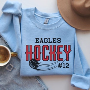 K&ouml;nnte beinhalten: Hellblauer Pullover mit dem Text "EAGLES HOCKEY #12" in roten und schwarzen Buchstaben. Eine schwarz-wei&szlig;e Grafik eines Hockeyschl&auml;gers befindet sich auf der linken Seite des Textes.