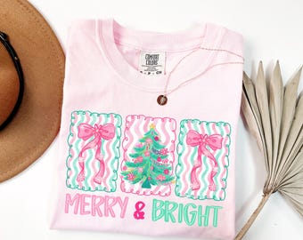 May include: A light pink Comfort Colors t-shirt with a Christmas-themed design. The design includes a Christmas tree and bows, with the words "MERRY & BRIGHT" in pink and green. A gold necklace is draped across the shirt.