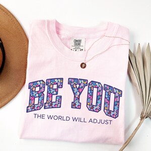 Pode incluir: Uma t-shirt de manga curta em rosa claro com decote redondo. A pe&ccedil;a apresenta o texto BE YOU em letras grandes de bloco preenchidas com um padr&atilde;o floral colorido, seguido pelo texto THE WORLD WILL ADJUST em letras menores de cor roxo escuro.