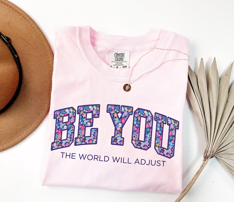 Pode incluir: Uma t-shirt de manga curta em rosa claro com decote redondo. A pe&ccedil;a apresenta o texto BE YOU em letras grandes de bloco preenchidas com um padr&atilde;o floral colorido, seguido pelo texto THE WORLD WILL ADJUST em letras menores de cor roxo escuro.