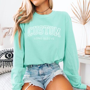 Op de afbeelding: Een mintgroen shirt met lange mouwen met het woord "CUSTOM" in witte blokletters. Onder het woord "CUSTOM" staat de tekst "LONG SLEEVE". Het shirt is gemaakt van een zacht materiaal en heeft een ronde hals.