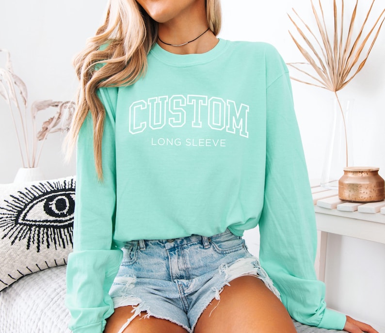 Op de afbeelding: Een mintgroen shirt met lange mouwen met het woord "CUSTOM" in witte blokletters. Onder het woord "CUSTOM" staat de tekst "LONG SLEEVE". Het shirt is gemaakt van een zacht materiaal en heeft een ronde hals.