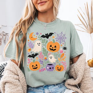 Może przedstawiać: Szałwiowo-zielony t-shirt z kr&oacute;tkim rękawem z kolorowym nadrukiem graficznym o tematyce Halloween. Wz&oacute;r zawiera pomarańczowe dynie, białe duszki, czarne nietoperze, fioletowy kociołek, pajęczynę, p&oacute;łksiężyc oraz cukierki.