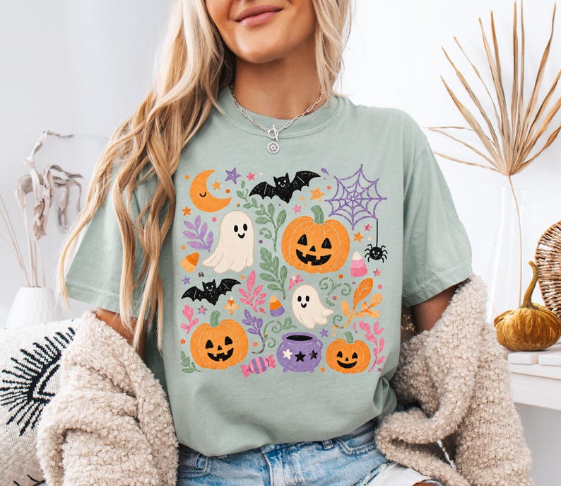 Może przedstawiać: Szałwiowo-zielony t-shirt z kr&oacute;tkim rękawem z kolorowym nadrukiem graficznym o tematyce Halloween. Wz&oacute;r zawiera pomarańczowe dynie, białe duszki, czarne nietoperze, fioletowy kociołek, pajęczynę, p&oacute;łksiężyc oraz cukierki.