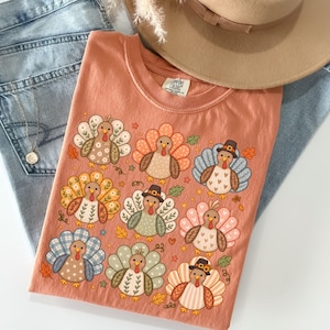 May include: A peach-colored t-shirt featuring a graphic print of nine stylized turkeys with various patterned feathers in shades of orange, blue, green, and white. Some turkeys wear small brown hats. The shirt has a crew neckline and a Comfort Colors label.