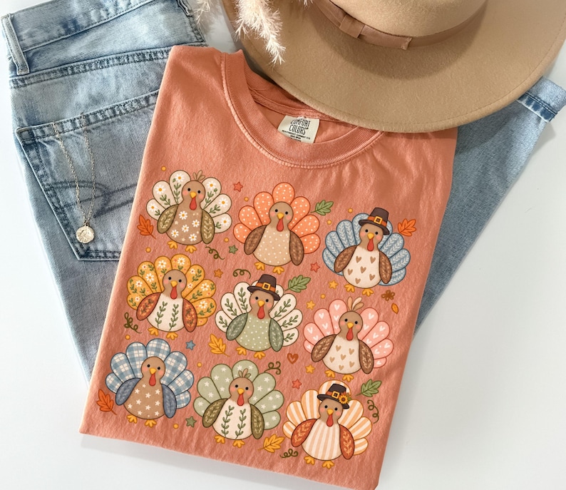 May include: A peach-colored t-shirt featuring a graphic print of nine stylized turkeys with various patterned feathers in shades of orange, blue, green, and white. Some turkeys wear small brown hats. The shirt has a crew neckline and a Comfort Colors label.