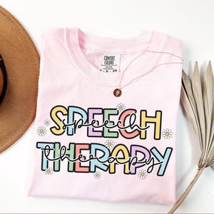 May include: A light pink t-shirt with the words "SPEECH THERAPY" in a colorful, playful font. The text is outlined in black and decorated with small daisy flowers. A gold necklace with a pendant rests on the shirt.