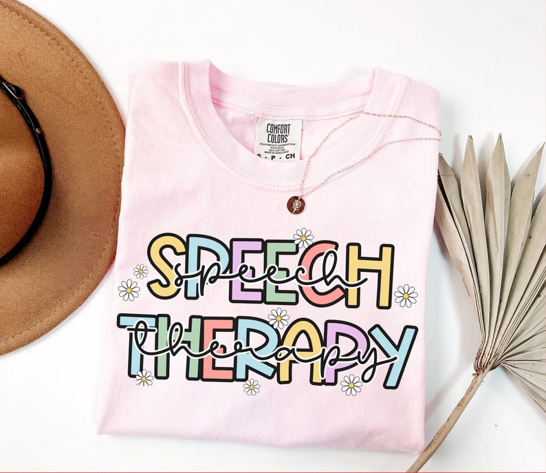 May include: A light pink t-shirt with the words "SPEECH THERAPY" in a colorful, playful font. The text is outlined in black and decorated with small daisy flowers. A gold necklace with a pendant rests on the shirt.