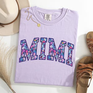 Puede incluir: Camiseta lila con la palabra "MIMI" en un dise&ntilde;o floral. La camiseta se presenta con un sombrero de paja, un collar dorado y sandalias marrones. La camiseta tiene una etiqueta Comfort Colors. Tema: ropa casual.