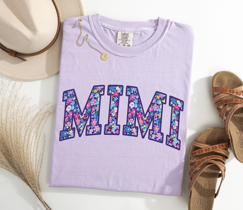 Puede incluir: Camiseta lila con la palabra "MIMI" en un dise&ntilde;o floral. La camiseta se presenta con un sombrero de paja, un collar dorado y sandalias marrones. La camiseta tiene una etiqueta Comfort Colors. Tema: ropa casual.