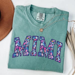 Puede incluir: Una camiseta Comfort Colors verde azulado con la palabra "MIMI" en letras grandes y florales. Un collar dorado con un peque&ntilde;o colgante descansa sobre la camisa. La camisa est&aacute; doblada, con un sombrero marr&oacute;n y una planta seca en el fondo.