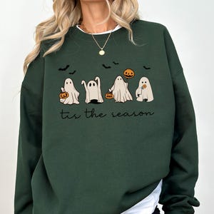 May include: Dark green sweatshirt featuring a Halloween design. The graphic includes four cartoon ghosts holding pumpkins and a pumpkin, with bats flying above. The text "tis the season" is below the ghosts.
