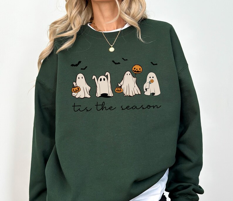 May include: Dark green sweatshirt featuring a Halloween design. The graphic includes four cartoon ghosts holding pumpkins and a pumpkin, with bats flying above. The text "tis the season" is below the ghosts.