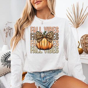 May include: White long-sleeved top featuring a pumpkin graphic and the words "FALL VIBES". The pumpkin has a leopard print bow. The shirt is a fall-themed design.