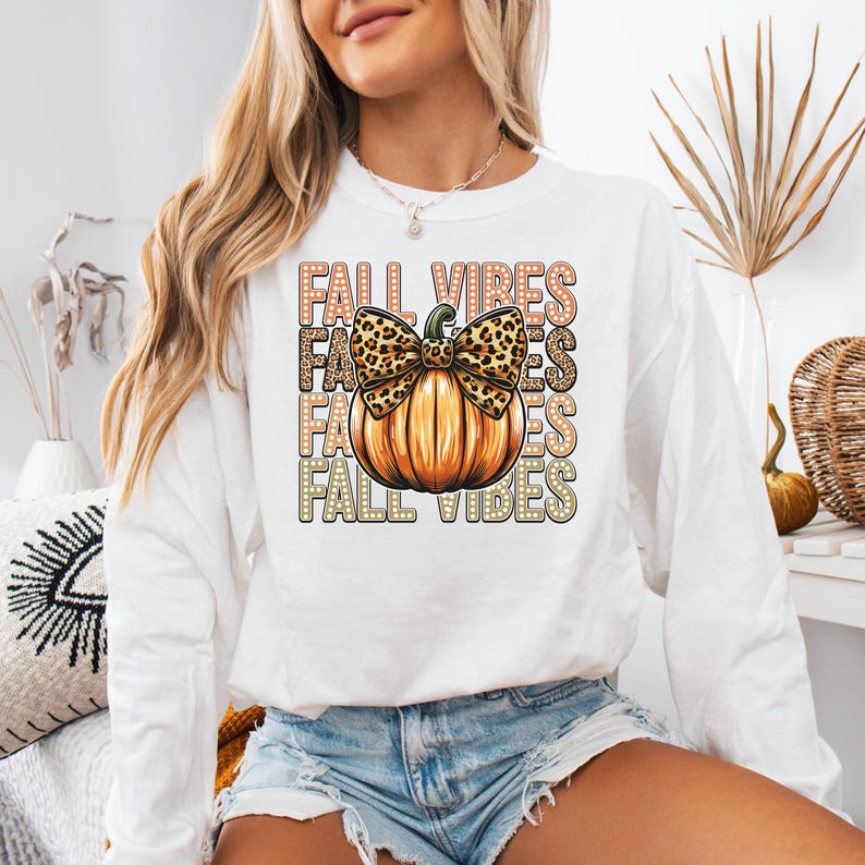 May include: White long-sleeved top featuring a pumpkin graphic and the words "FALL VIBES". The pumpkin has a leopard print bow. The shirt is a fall-themed design.
