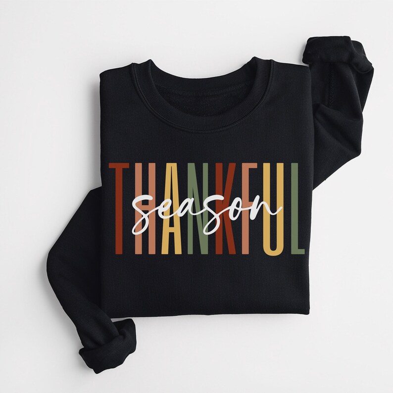 Puede incluir: Sudadera negra con el texto "Thankful Season" en rayas verticales de colores. El texto est&aacute; en un estilo de fuente retro.
