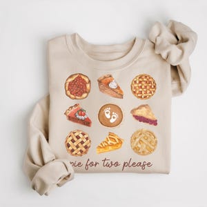 May include: A beige sweatshirt with a graphic of various pies, including cherry, pumpkin, pecan, and apple. The text "pie for two please" is printed below the pies.