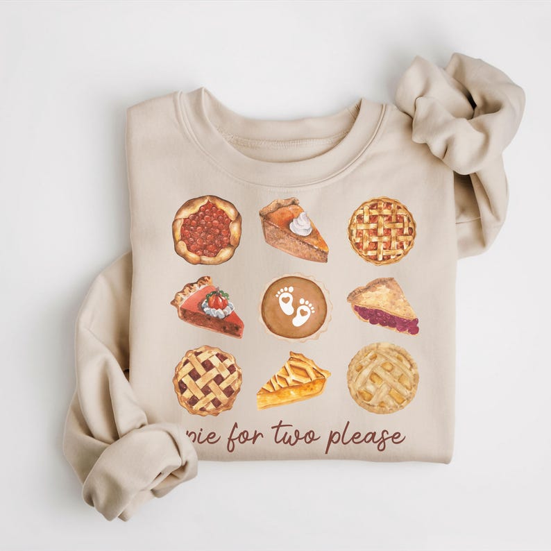 May include: A beige sweatshirt with a graphic of various pies, including cherry, pumpkin, pecan, and apple. The text "pie for two please" is printed below the pies.