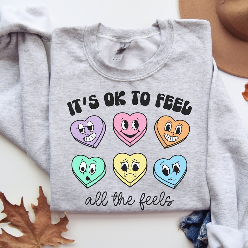 Pu&ograve; includere: Una felpa grigia con la scritta "It's OK to feel all the feels" stampata su di essa. Il testo &egrave; in un font retr&ograve; ed &egrave; circondato da sei cuori di caramelle colorati con diverse espressioni facciali.