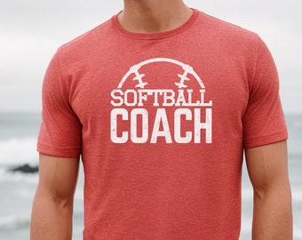 May include: Red t-shirt with the words "SOFTBALL COACH" in white, set within a softball design. The shirt is short-sleeved and has a textured appearance. The background is a blurred view of the ocean.