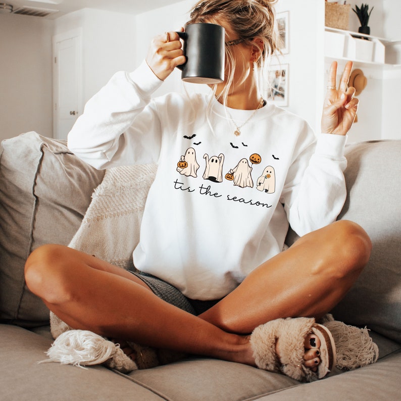 May include: White sweatshirt featuring a Halloween-themed design with cartoon ghosts, pumpkins, and bats. The phrase "tis the season" is printed below the graphic. The sweatshirt is worn by a person sitting on a couch.