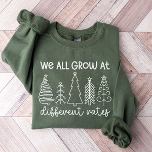 May include: A green sweatshirt with the text "We all grow at different rates" and a row of five stylized Christmas trees printed in white.