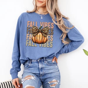 May include: A blue long-sleeved top with a pumpkin design and the words "FALL VIBES". The pumpkin has a leopard print bow. The pumpkin is orange with brown shading.