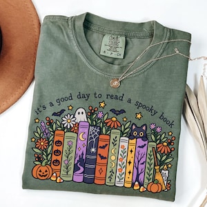 May include: An olive green t-shirt featuring a graphic print of colourful books, flowers, a ghost, a black cat, and pumpkins. The text reads, it's a good day to read a spooky book. The shirt has a Comfort Colors brand label at the inner neckline.