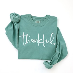 May include: A teal long-sleeved shirt with the word "thankful" in white script across the front. A small white heart is next to the word. The shirt has a crew neck and rolled-up sleeves.