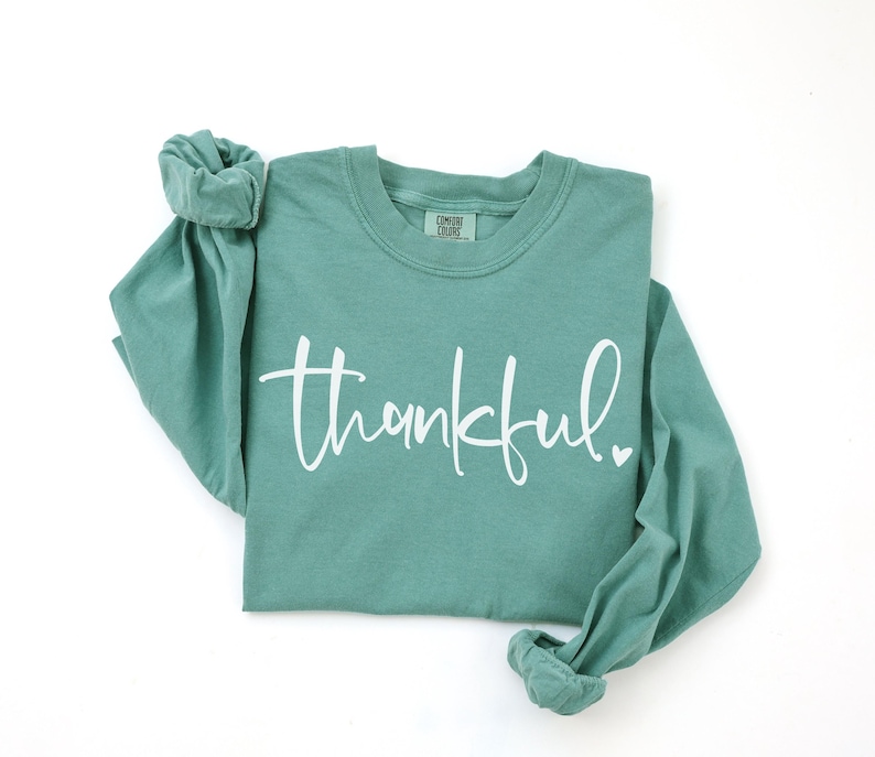 May include: A teal long-sleeved shirt with the word "thankful" in white script across the front. A small white heart is next to the word. The shirt has a crew neck and rolled-up sleeves.