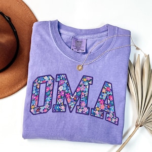 May include: A lilac Comfort Colors t-shirt with the word "OMA" in large, floral-patterned letters. A gold necklace and a brown hat are also in the image. The shirt is folded and has a small tag.
