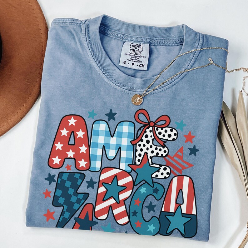 May include: A pale blue Comfort Colors t-shirt featuring the word "AMERICA" in a patriotic design. The letters are red, white, and blue with star and stripe patterns. A gold necklace with a pendant is also visible.