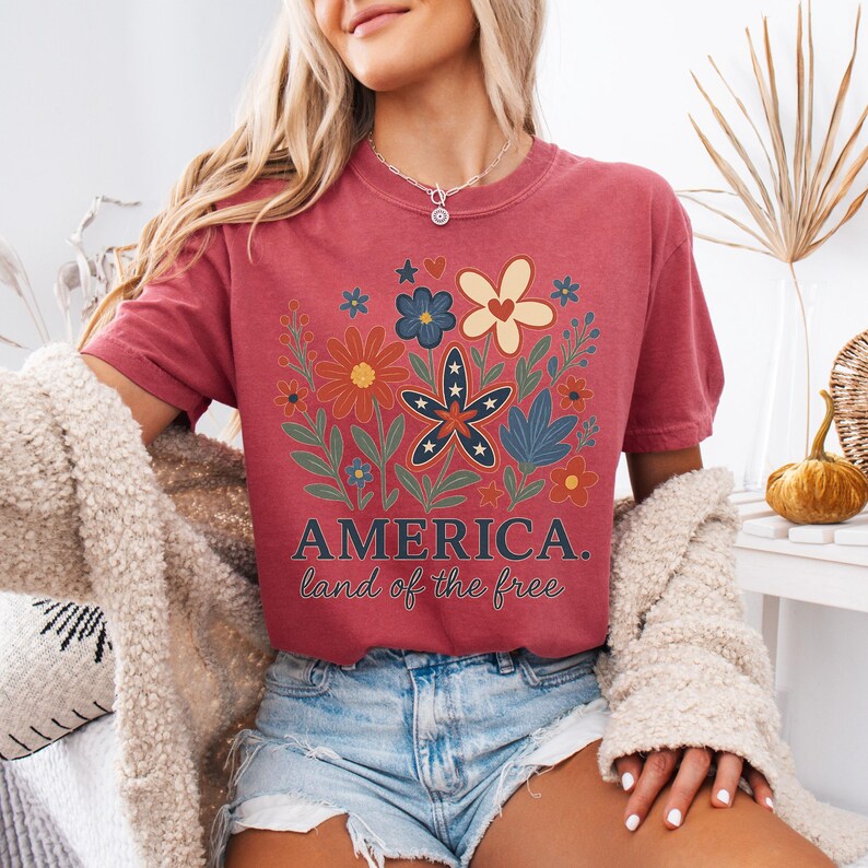 Peut inclure: Un t-shirt rouge avec un motif floral comportant des &eacute;toiles et des rayures. Le texte "AMERICA. land of the free" est imprim&eacute; sur le t-shirt.