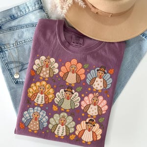 May include: A plum-colored t-shirt featuring a graphic print of nine stylized turkeys with various patterned feathers, including floral, plaid, and heart designs. Some turkeys wear small brown hats. The shirt is displayed with light blue jeans and a tan hat.