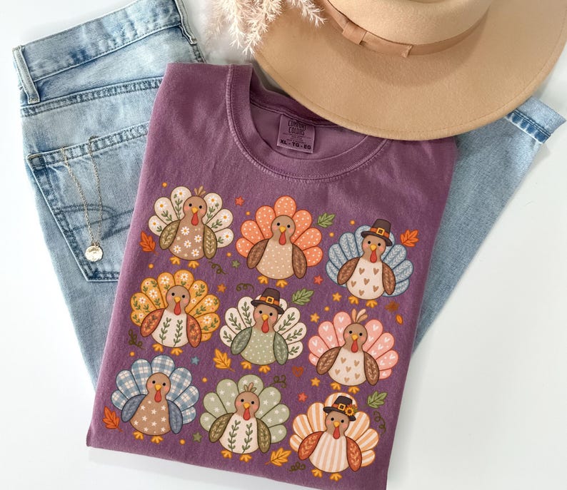 May include: A plum-colored t-shirt featuring a graphic print of nine stylized turkeys with various patterned feathers, including floral, plaid, and heart designs. Some turkeys wear small brown hats. The shirt is displayed with light blue jeans and a tan hat.