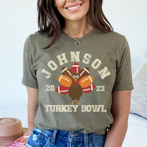 Op de afbeelding: Een groen T-shirt met een vintage-stijl afbeelding van een kalkoen die voetballen als veren draagt. De tekst "JOHNSON 20 23 TURKEY BOWL" is op het shirt gedrukt.