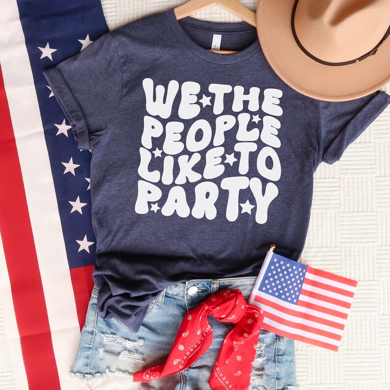 May include: A navy blue t-shirt with white text that reads "WE THE PEOPLE LIKE TO PARTY" with stars around the words. The shirt is laid out on a white surface with an American flag and a brown cowboy hat.