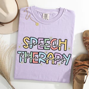 May include: A light purple t-shirt with the words "SPEECH THERAPY" in colorful block letters and daisy accents. A gold necklace and a beige hat are next to the shirt, along with brown sandals.