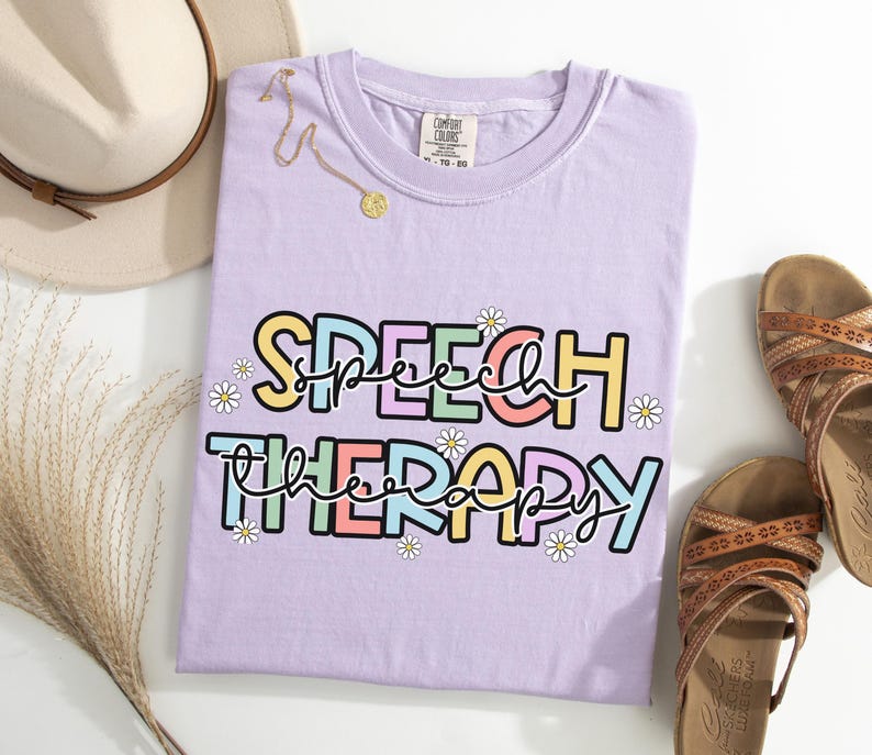 May include: A light purple t-shirt with the words "SPEECH THERAPY" in colorful block letters and daisy accents. A gold necklace and a beige hat are next to the shirt, along with brown sandals.